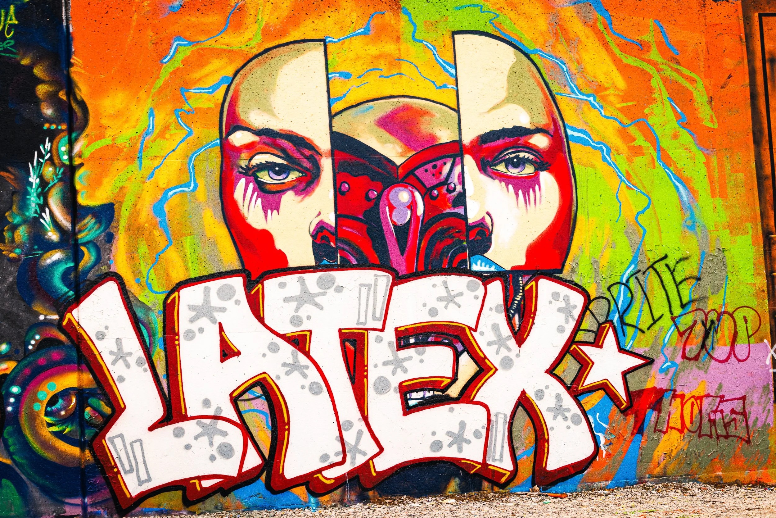 Colorful graffiti mural depicting a stylized face with bright eyes, surrounded by vibrant abstract patterns and chaotic lines on a wall.