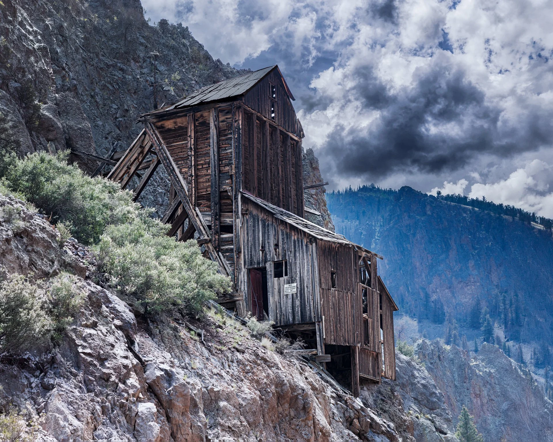 An old, weathered wooden mine shaft building built into the side of a rocky mountain with a cloudy sky overhead.