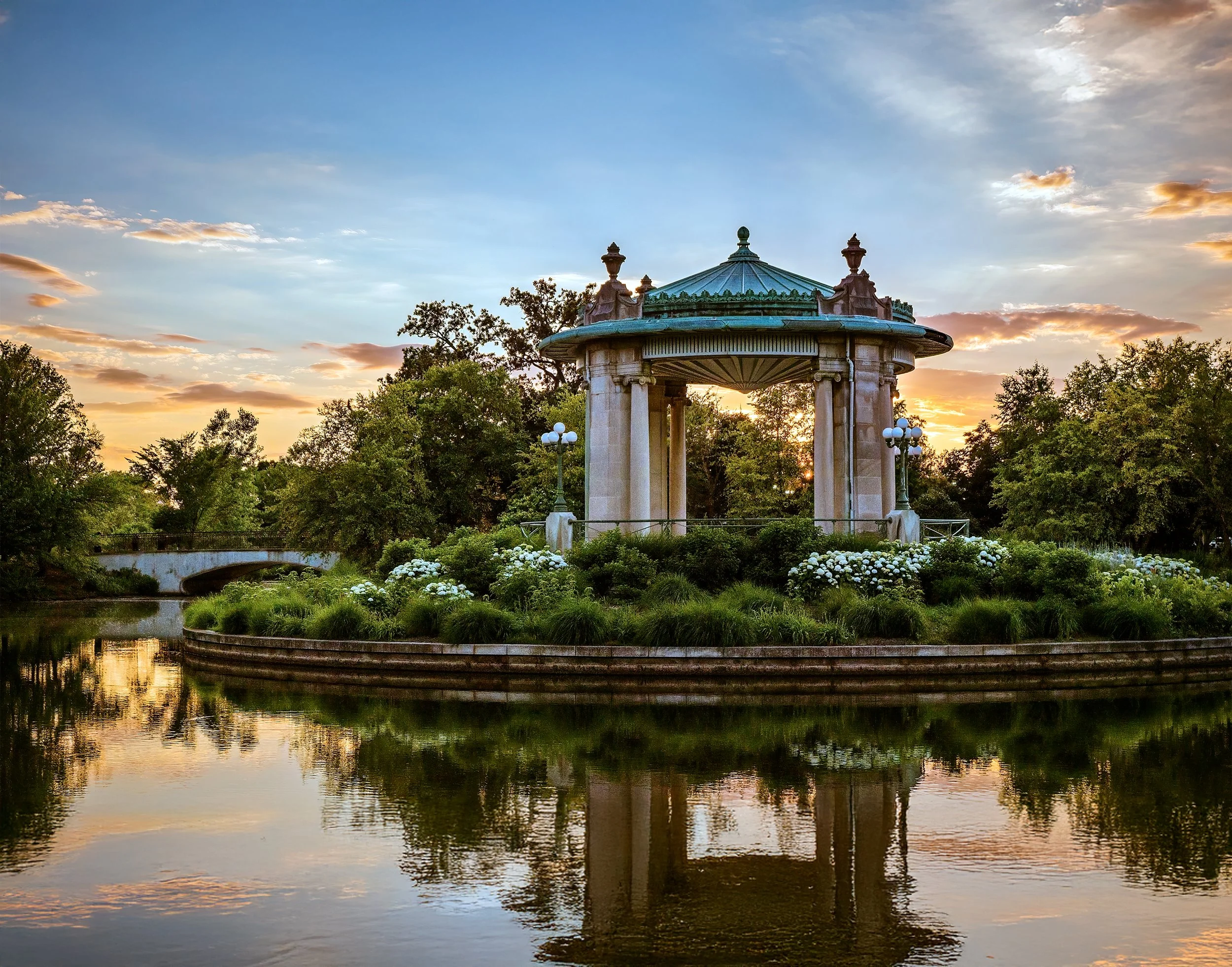 A historic gazebo with a green roof and ornate details, situated by a river at sunset, surrounded by trees and white flowers.