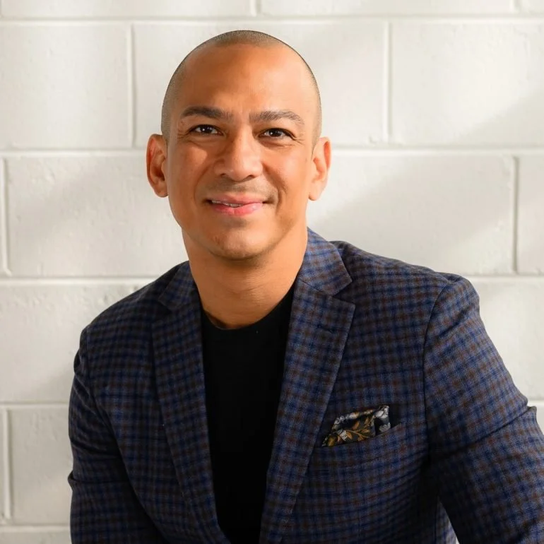 Portrait of a smiling man with a shaved head, wearing a dark checkered blazer and black shirt, against a white brick wall background.