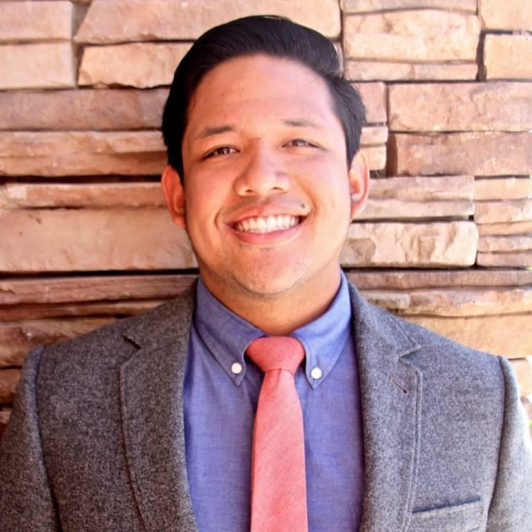 Portrait of a smiling man wearing a suit, light blue shirt, and pink tie, standing against a stone wall background.