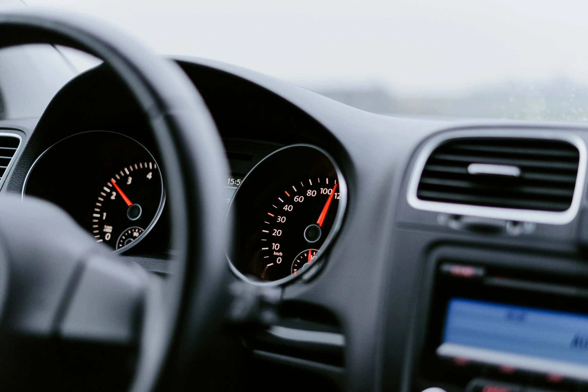 View of a car dashboard through the steering wheel, with speedometer and tachometer, on a rainy day with foggy windshield.