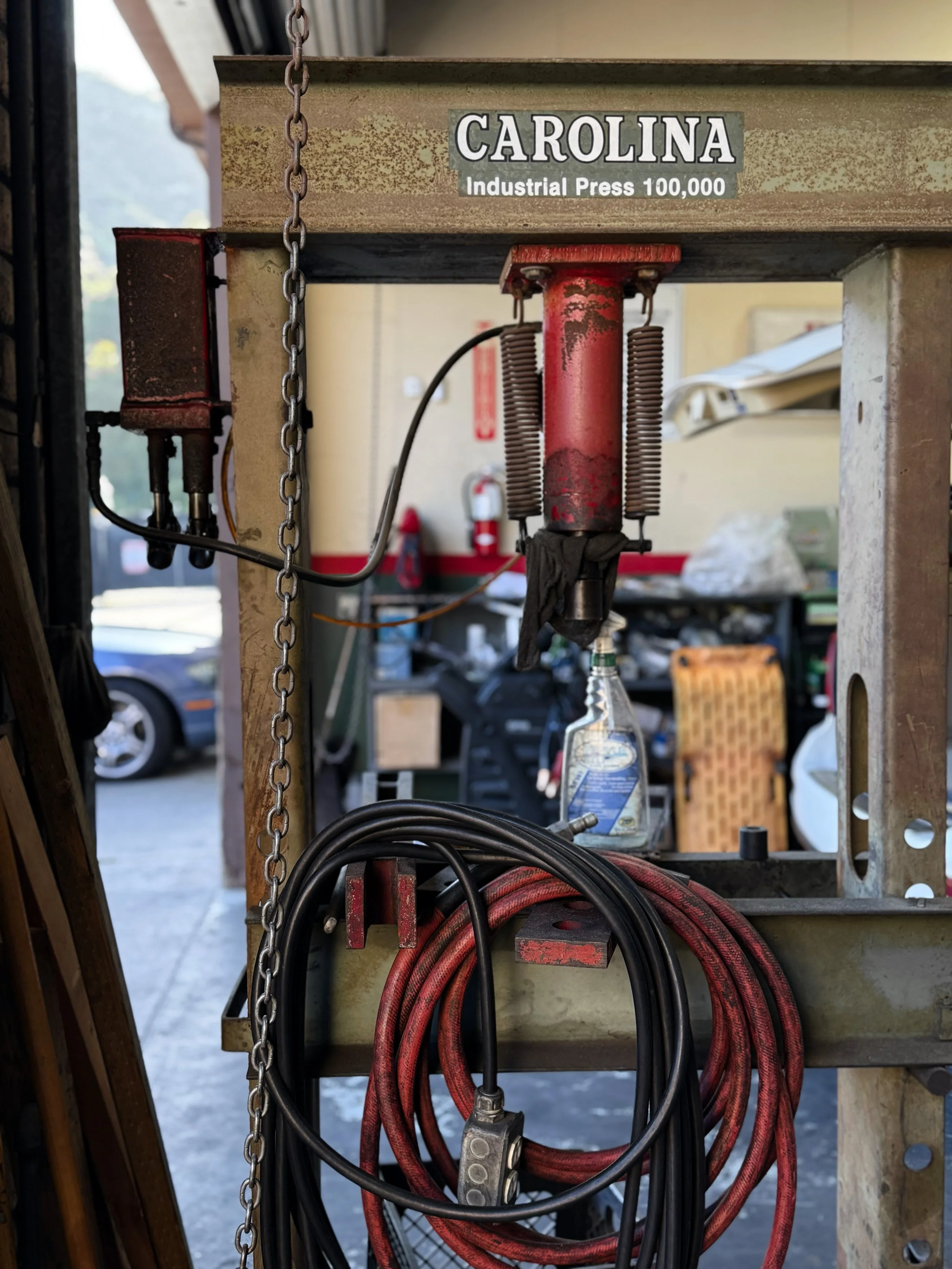 A close-up view of a metal valve and hoses attached to an industrial press labeled 'Carolina' with a capacity of 100,000 pounds.