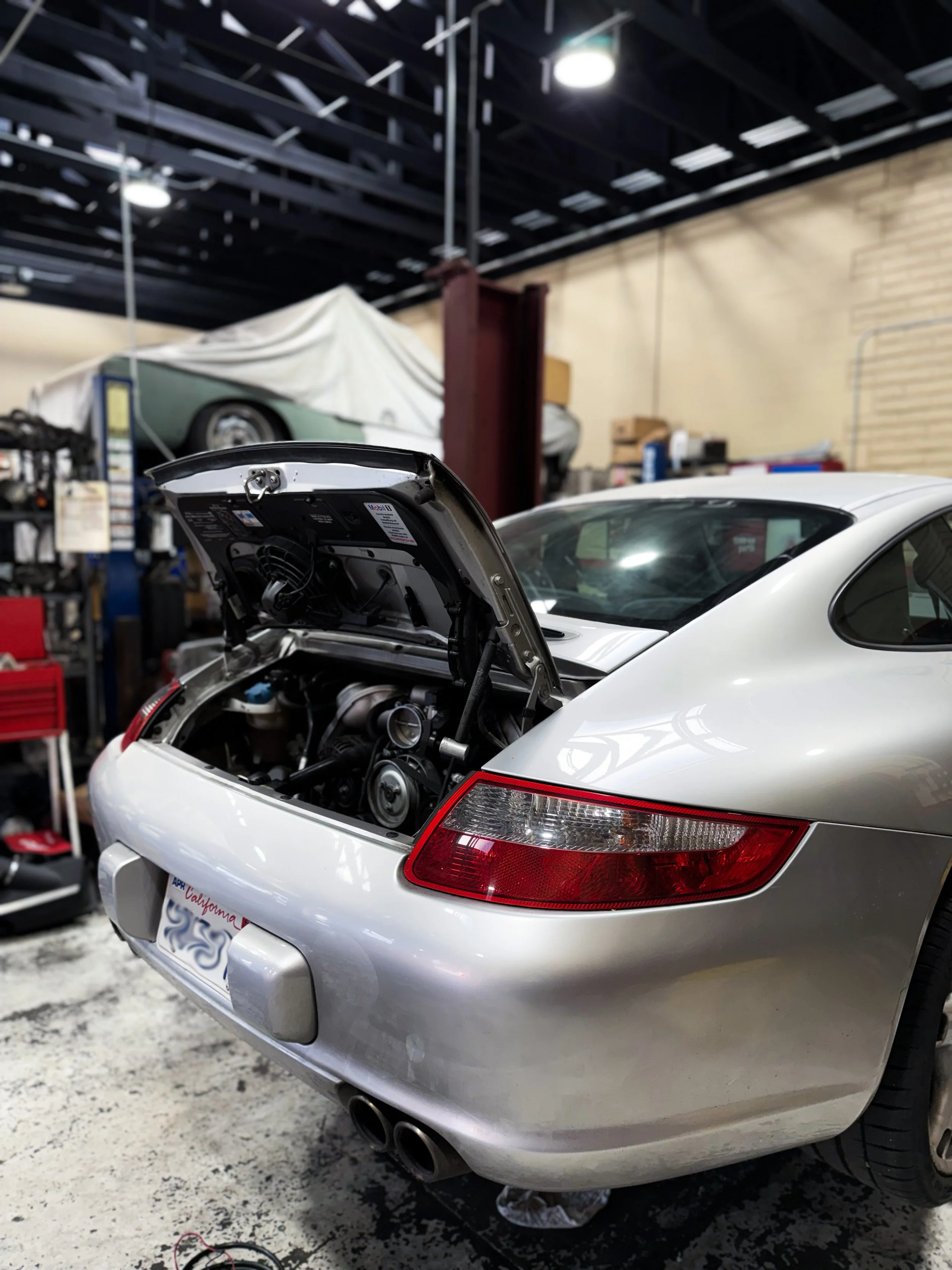 A silver Porsche sports car with its rear hood open in a garage or workshop. The car has a California license plate and dual exhaust pipes. There is a red tool chest and shelves with various tools and car parts in the background.