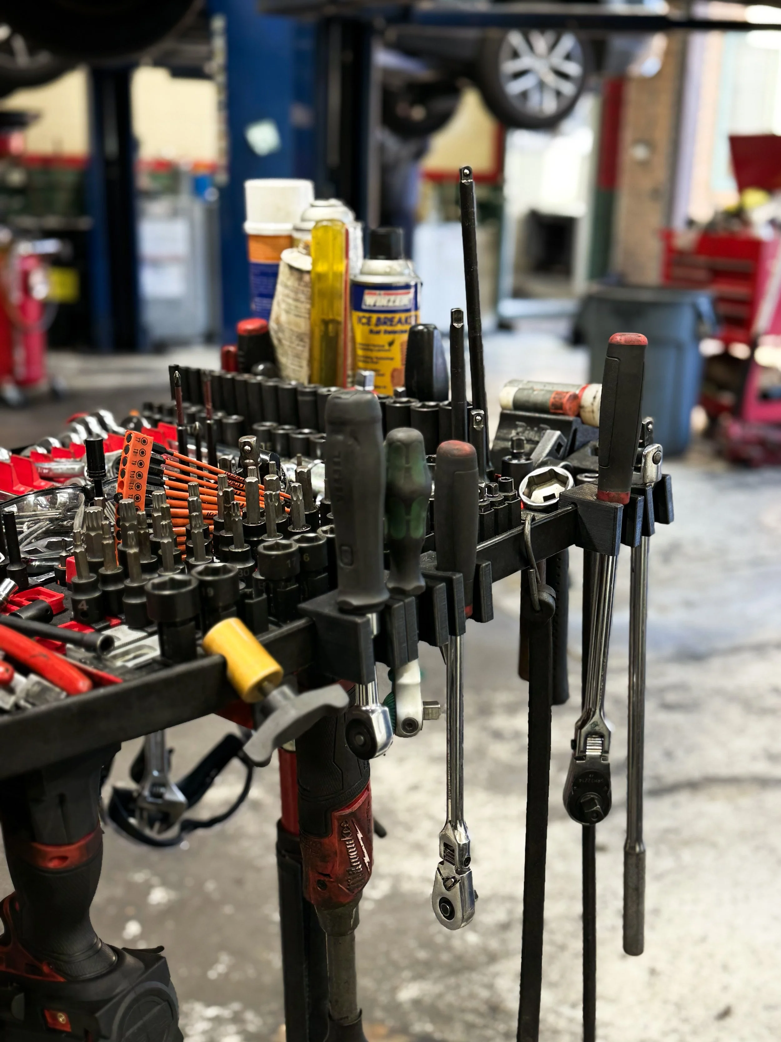 Tool cart in a garage with various hand tools, spark plugs, and bottles of lubricant, with tires and shelves in the background.