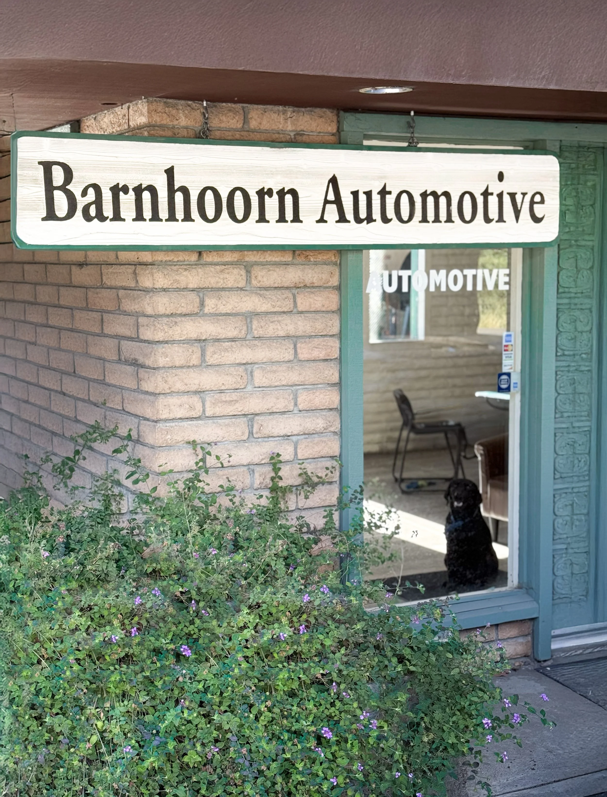 Sign for Barnhoorn Automotive outside a brick building with a green door. A black dog sits on the porch near the door. There are purple flowers and green bushes in the foreground.