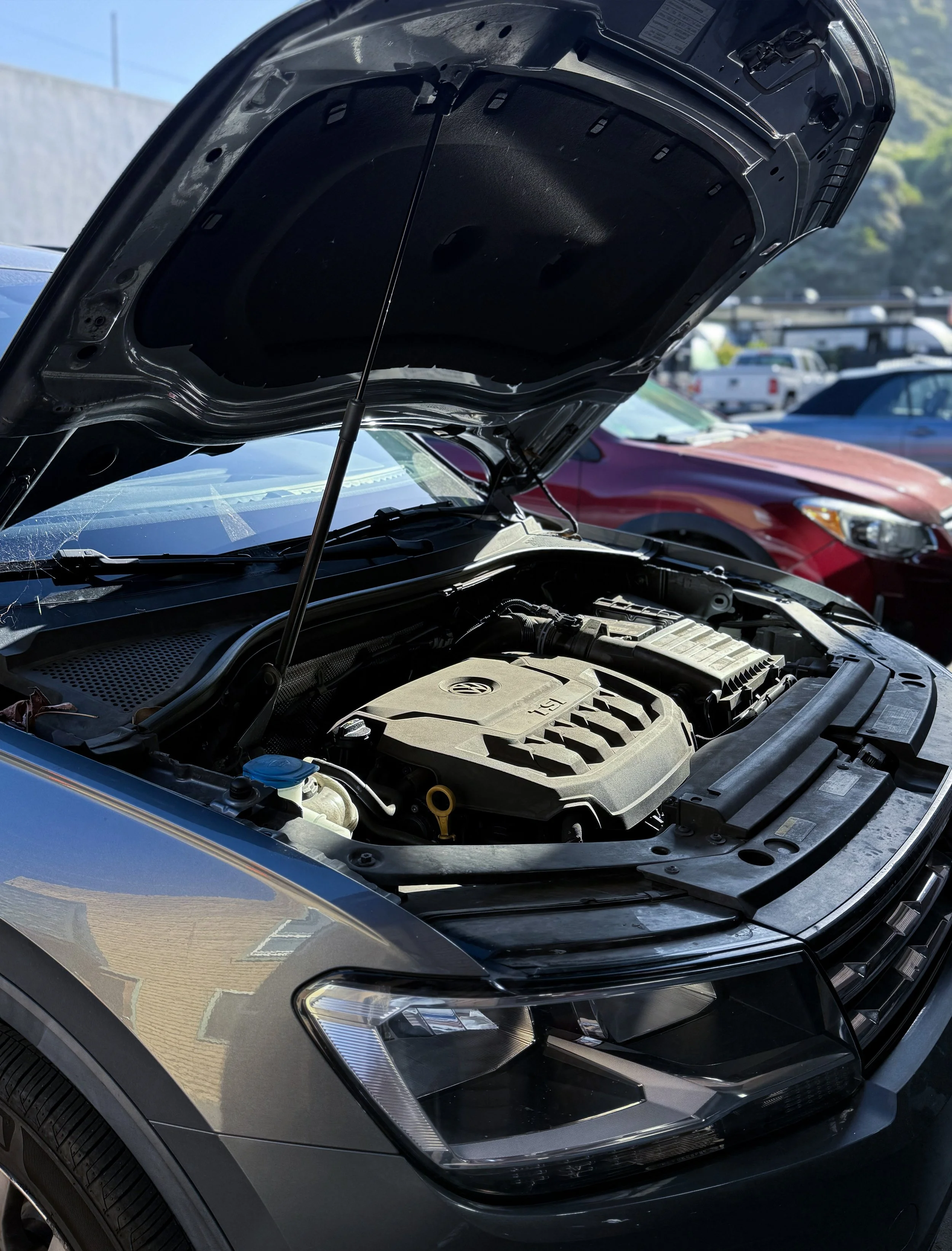 Close-up of a car with the hood open, showing the engine compartment, parked outdoors in a lot with other vehicles. The front of the car is black with a shiny finish, and the headlight is visible.