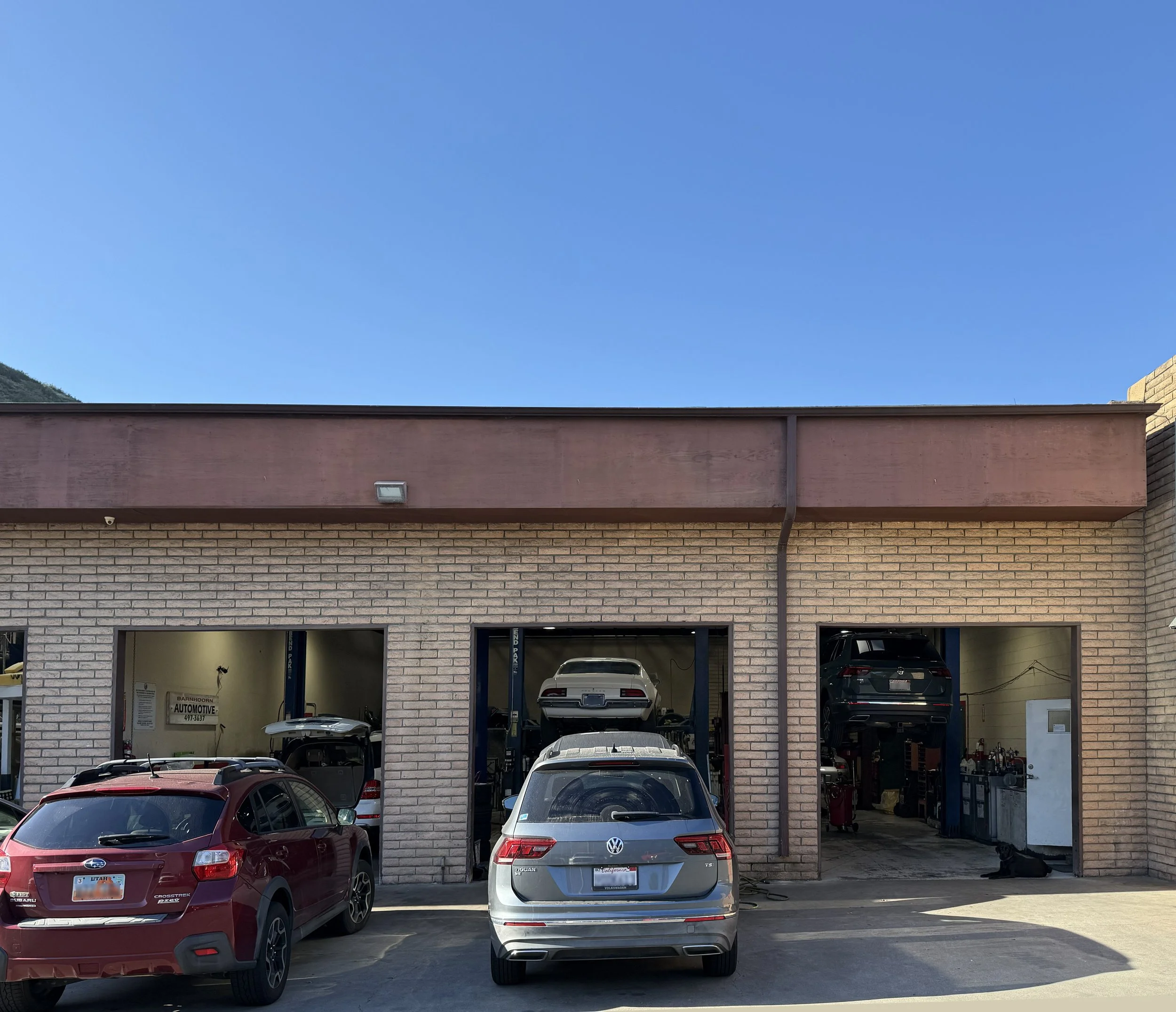 A parking lot in front of a brick building with an automotive repair shop. Several cars are parked outside, and some are inside the garage bays. The building has a beige brick exterior with a dark brown trim, and there is a sign inside that indicates it is an automotive business.
