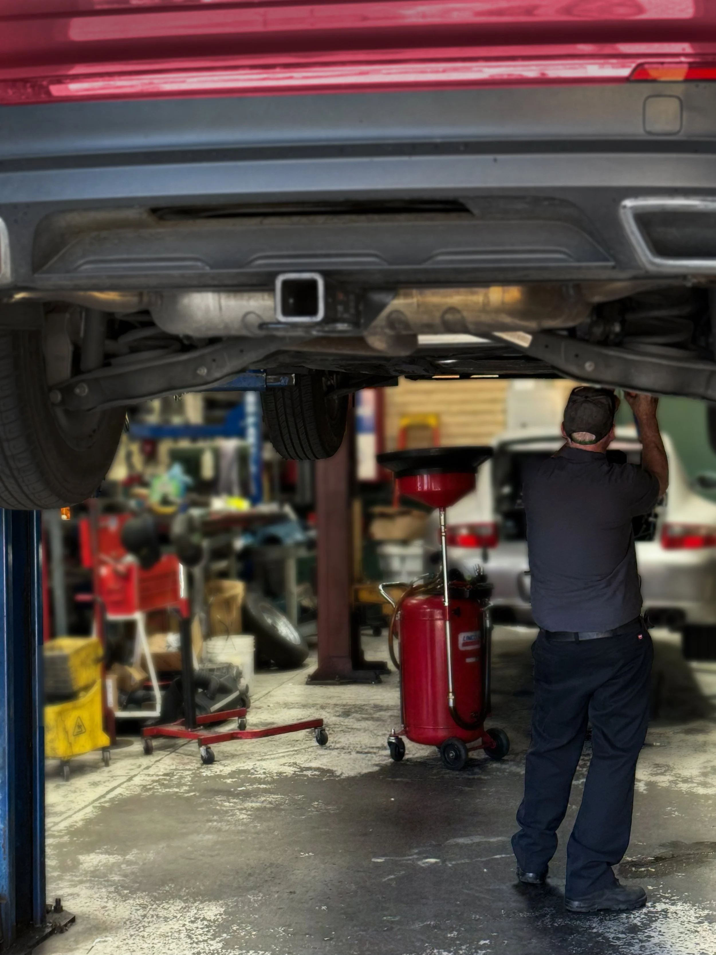 Auto mechanic working underneath a vehicle lifted on a hydraulic lift in an auto repair shop, with tools and equipment in the background.
