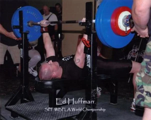 Ed Huffman winning the Southern Powerlifting Federation World Championship. Ed Huffman lying on a bench, lifting a barbell with blue and red weights, during a powerlifting competition. Several spectators and officials are nearby.
