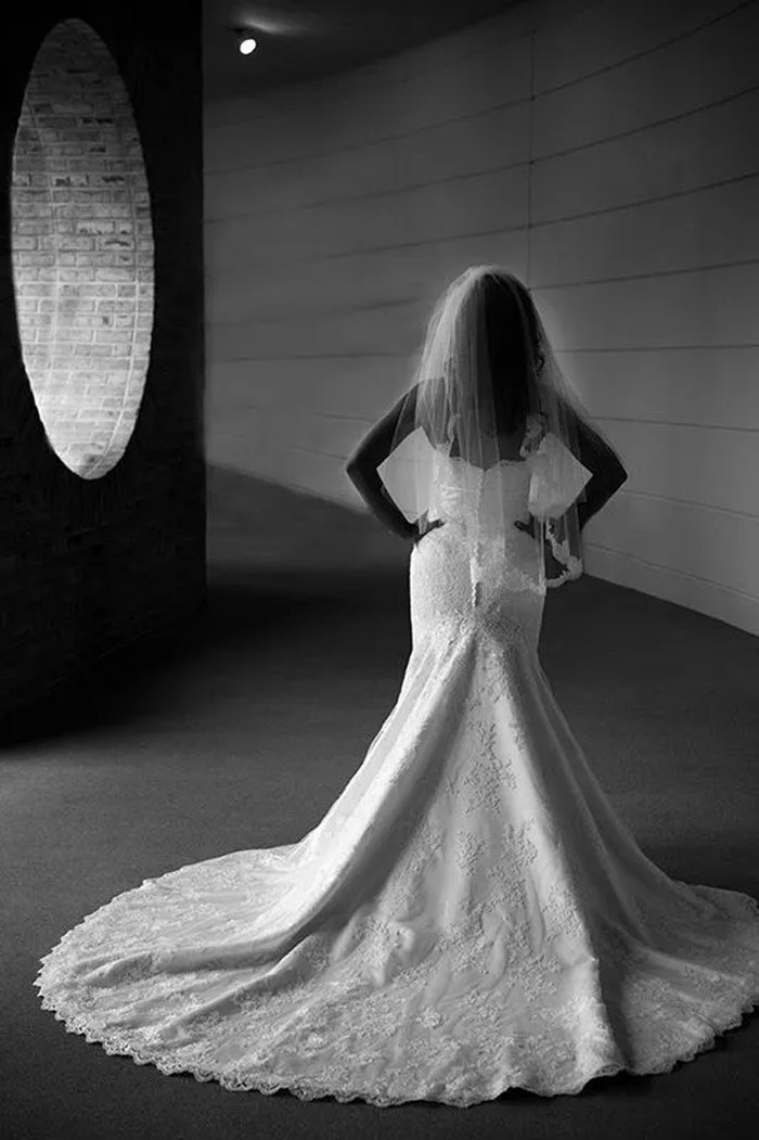 A bride in a wedding gown with a long train, standing with her hands on her hips, wearing a veil, in a dimly lit hallway with a brick wall and circular window.