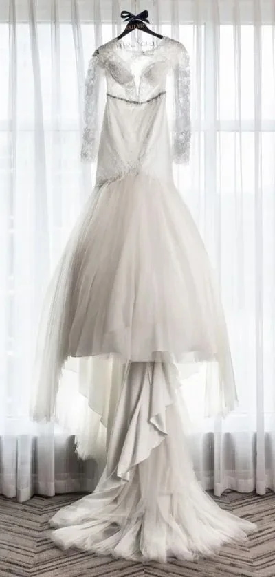 A white wedding dress hanging on a hanger in front of sheer white curtains, with a layered skirt, lace long sleeves, and a lace bodice.