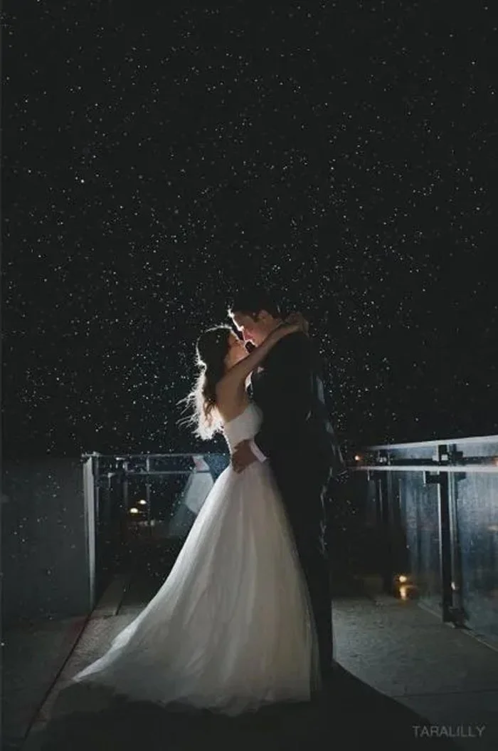 A bride and groom sharing a romantic kiss on a rooftop at night, under a starry sky, with rain falling.