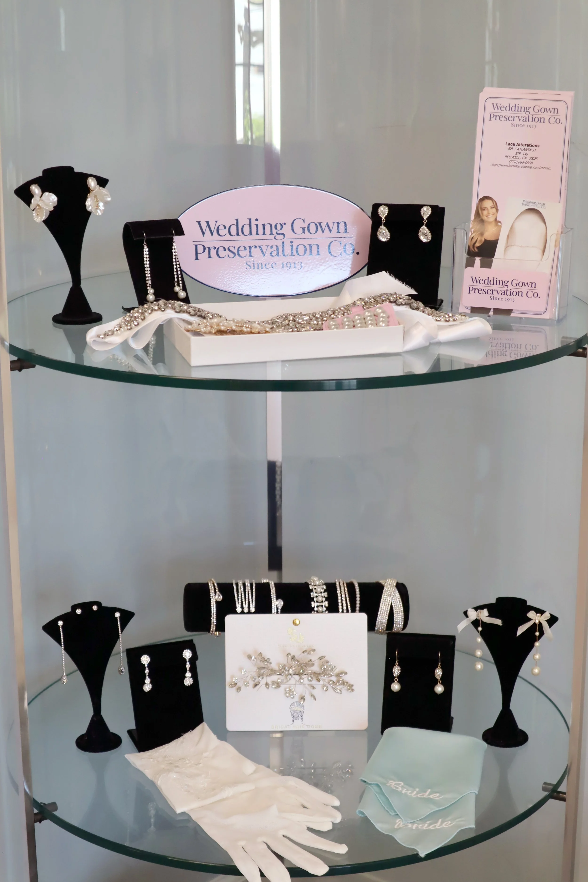 Display of bridal jewelry and accessories, including earrings, necklaces, and gloves, on black velvet stands and a glass shelf.