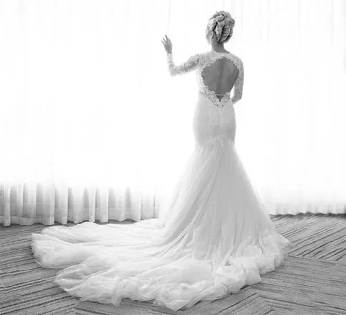 A woman in a wedding dress standing in front of a window with curtains, with her back to the camera and her hand raised, in a room with a patterned carpet.