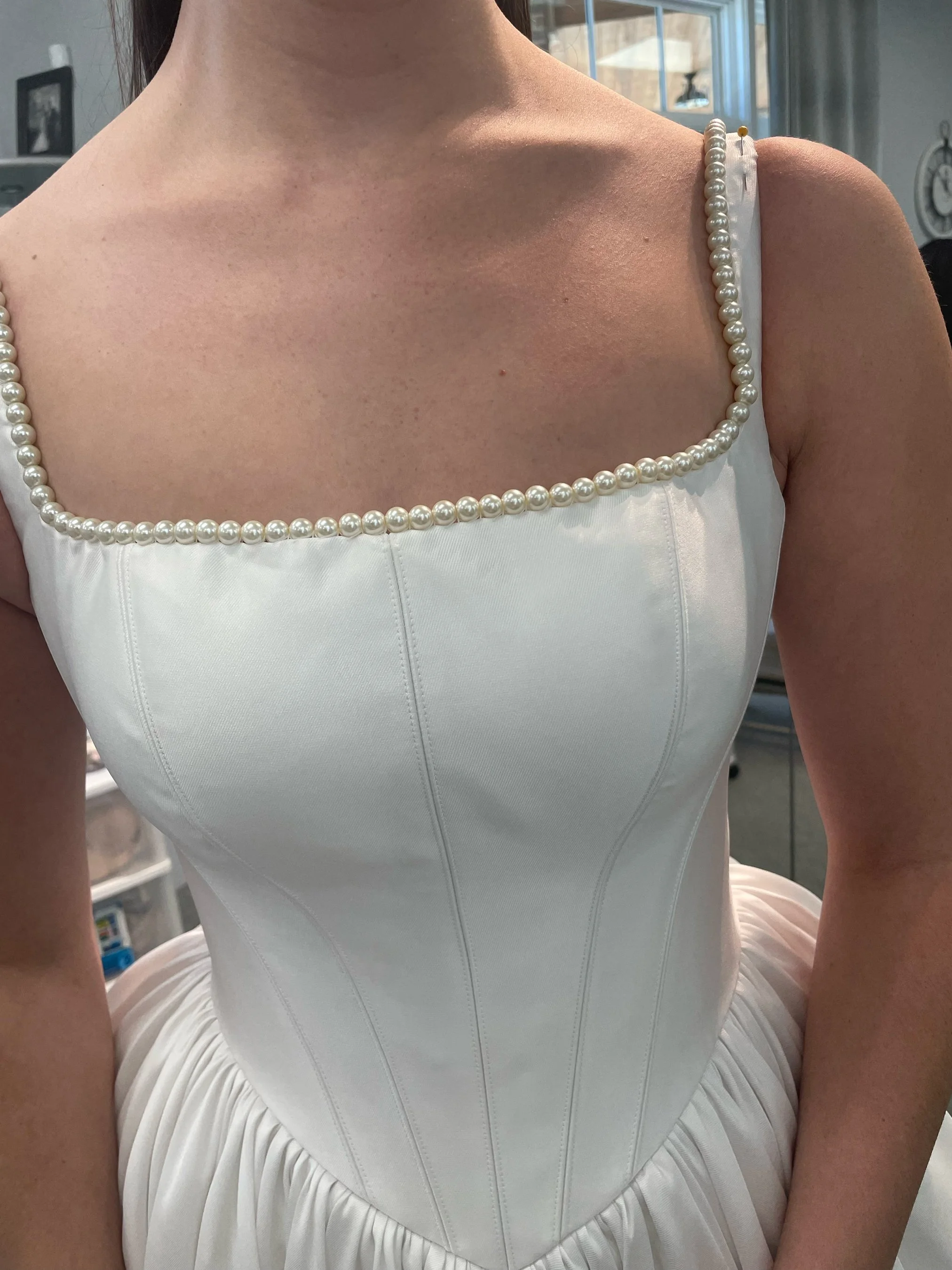 Close-up of a woman wearing a white dress with pearl straps, showing her shoulders and upper chest, with a background of windows and a clock.
