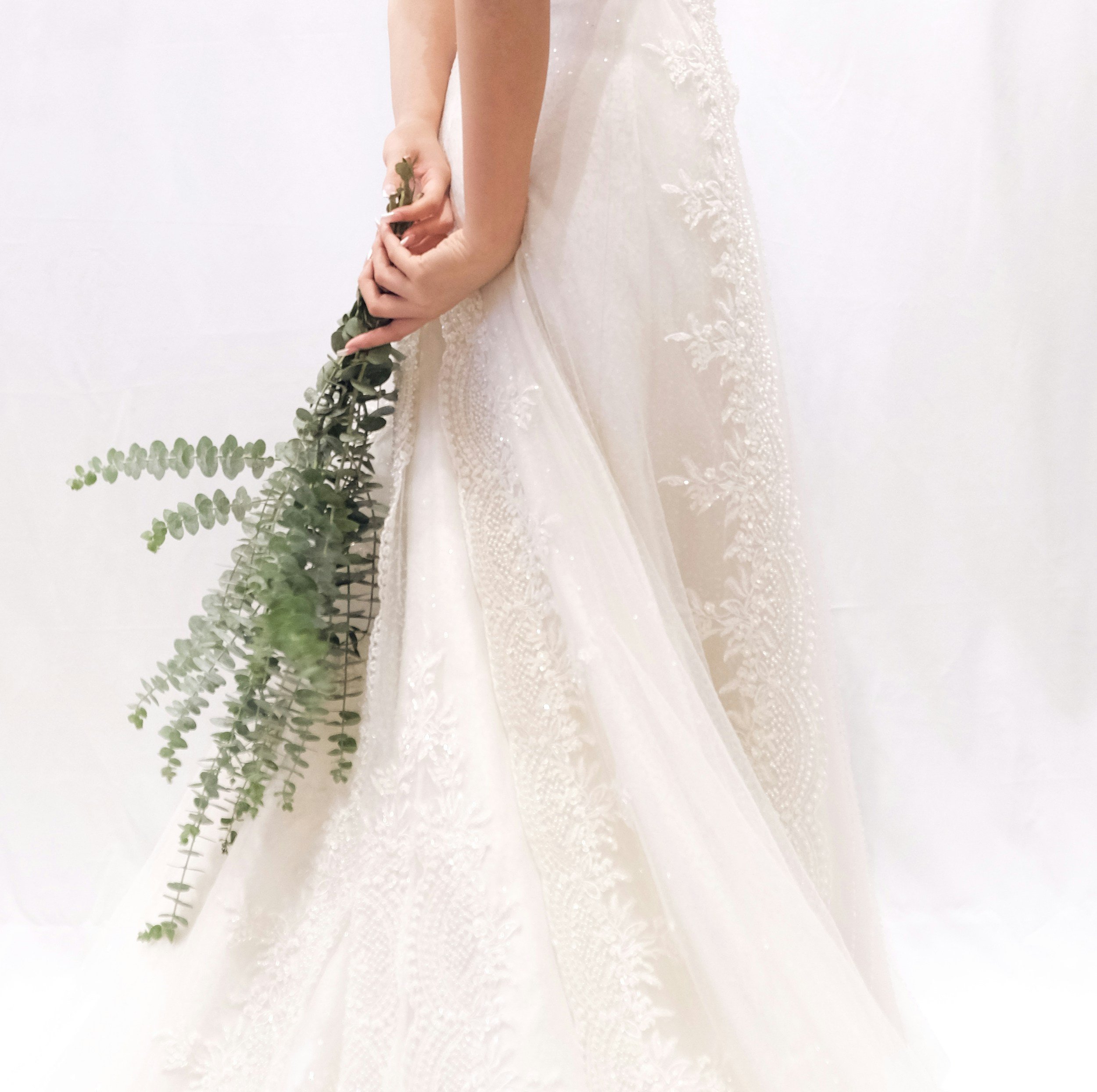 Close-up of a woman in a white lace wedding dress holding a sprig of eucalyptus leaves against a plain white background.