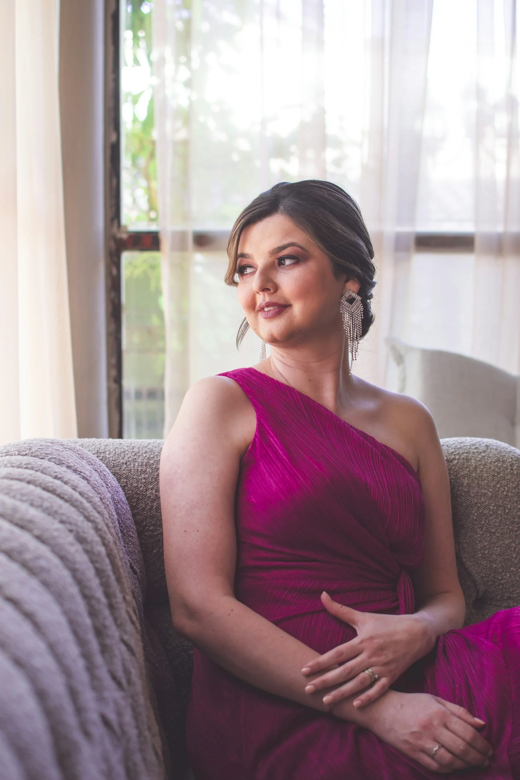A woman with dark hair in an elegant updo, wearing a magenta strapless dress and large dangling earrings, sitting on a couch near a window with sheer curtains, looking to the side with a gentle smile.