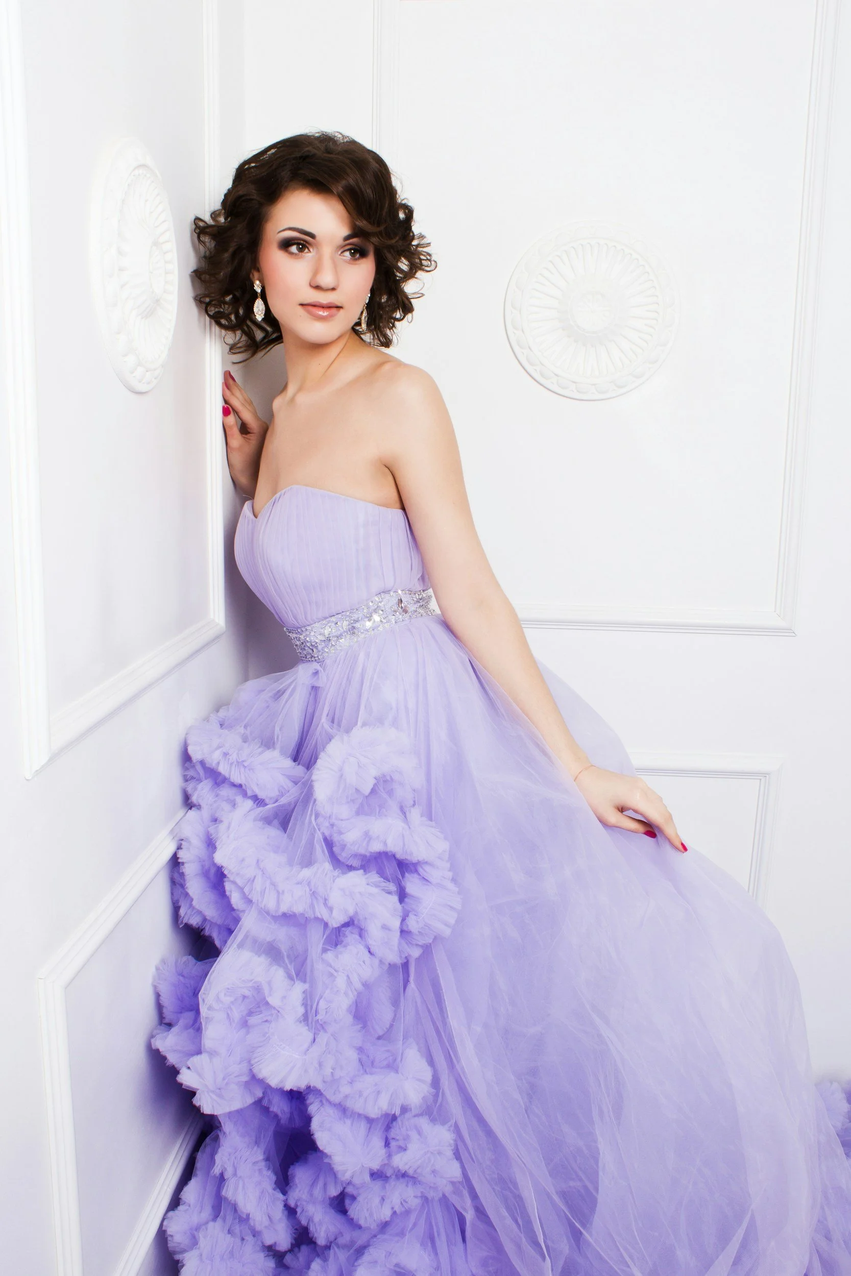 A young woman with dark, curly hair wearing a strapless lavender gown with ruffles and tulle, standing against a white paneled wall, looking to the side.