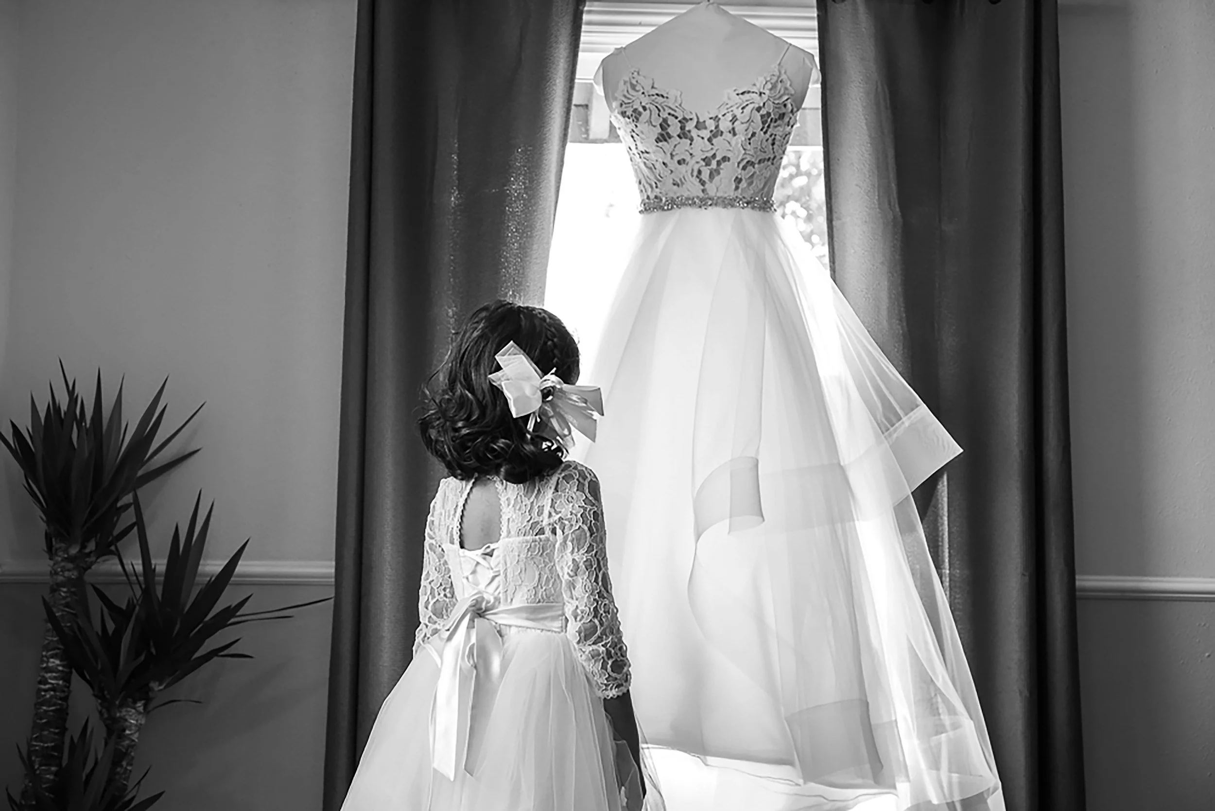 A young girl in a lace dress with a large bow in her hair standing in front of a wedding dress hanging in front of a window.