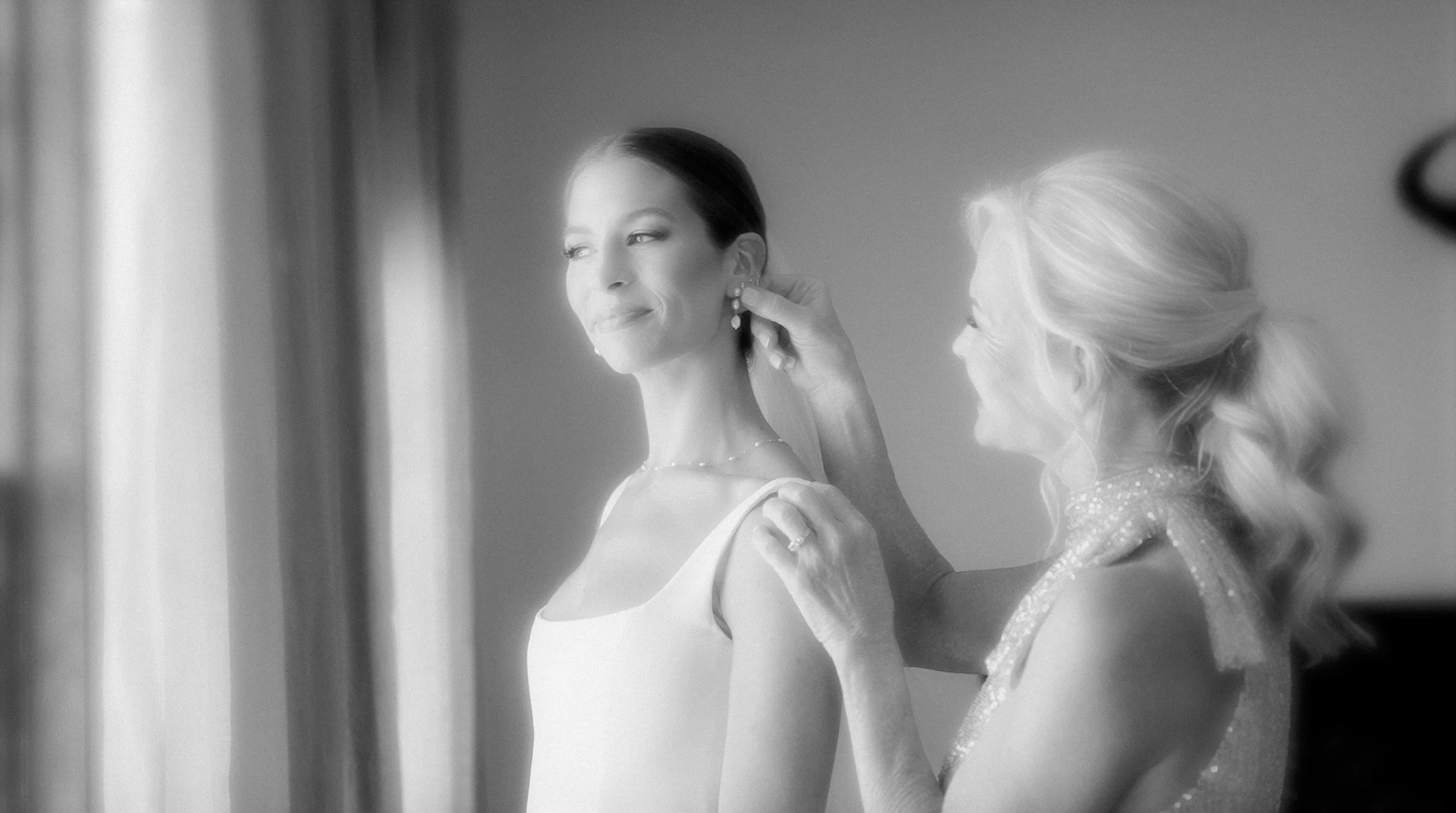 A woman in a gown having earrings adjusted by another woman, with a soft glow from a nearby window, in black and white.