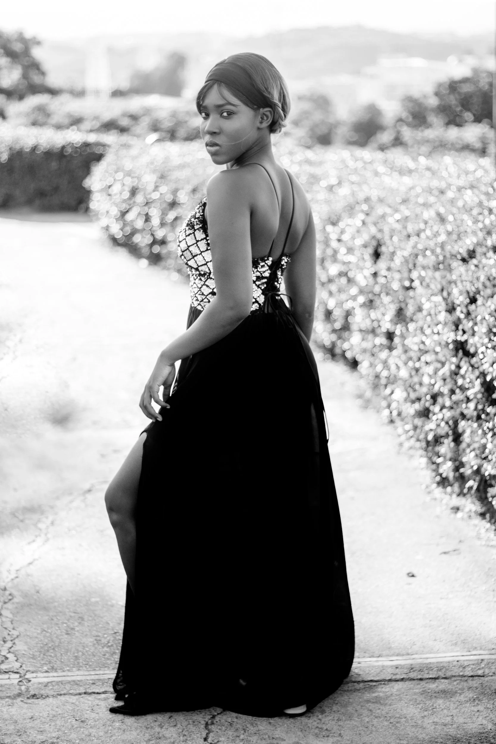Black and white photo of a woman with short hair wearing a dress with thin straps, standing outdoors on a concrete surface, with bushes and a blurred background behind her.
