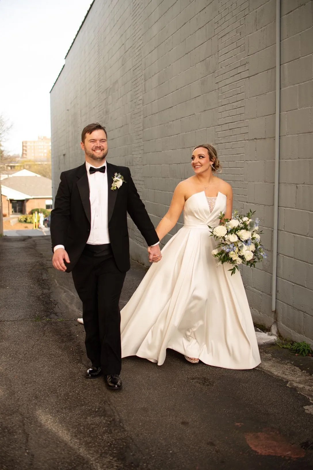 A bride and groom holding hands and walking outside near a gray brick wall, smiling. The bride is wearing a white wedding dress and holding a bouquet; the groom is in a black tuxedo with a bow tie.