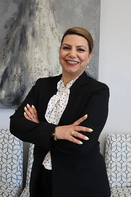A woman with short brown hair, wearing a black blazer and white polka dot blouse, smiling with arms crossed, standing in front of an abstract painting and patterned chairs.