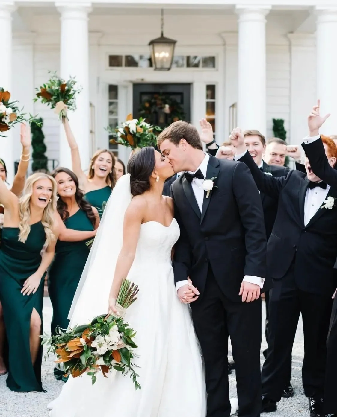 A wedding celebration with a bride and groom kissing, surrounded by joyful friends and family outside a white building with columns.
