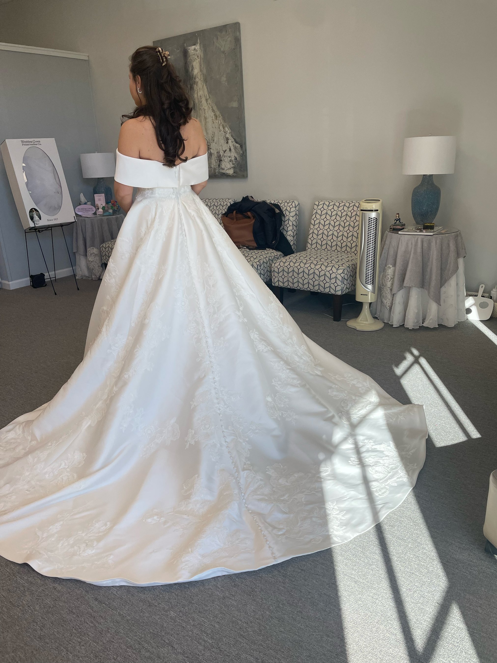 A woman in a white wedding gown standing in a room with gray walls, facing away from the camera, with sunlight casting shadows on the carpet.