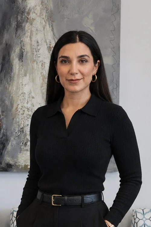 A woman with long dark hair, wearing a black long-sleeve shirt with a collar, gold hoop earrings, and a black belt, standing in front of an abstract painting on a wall.