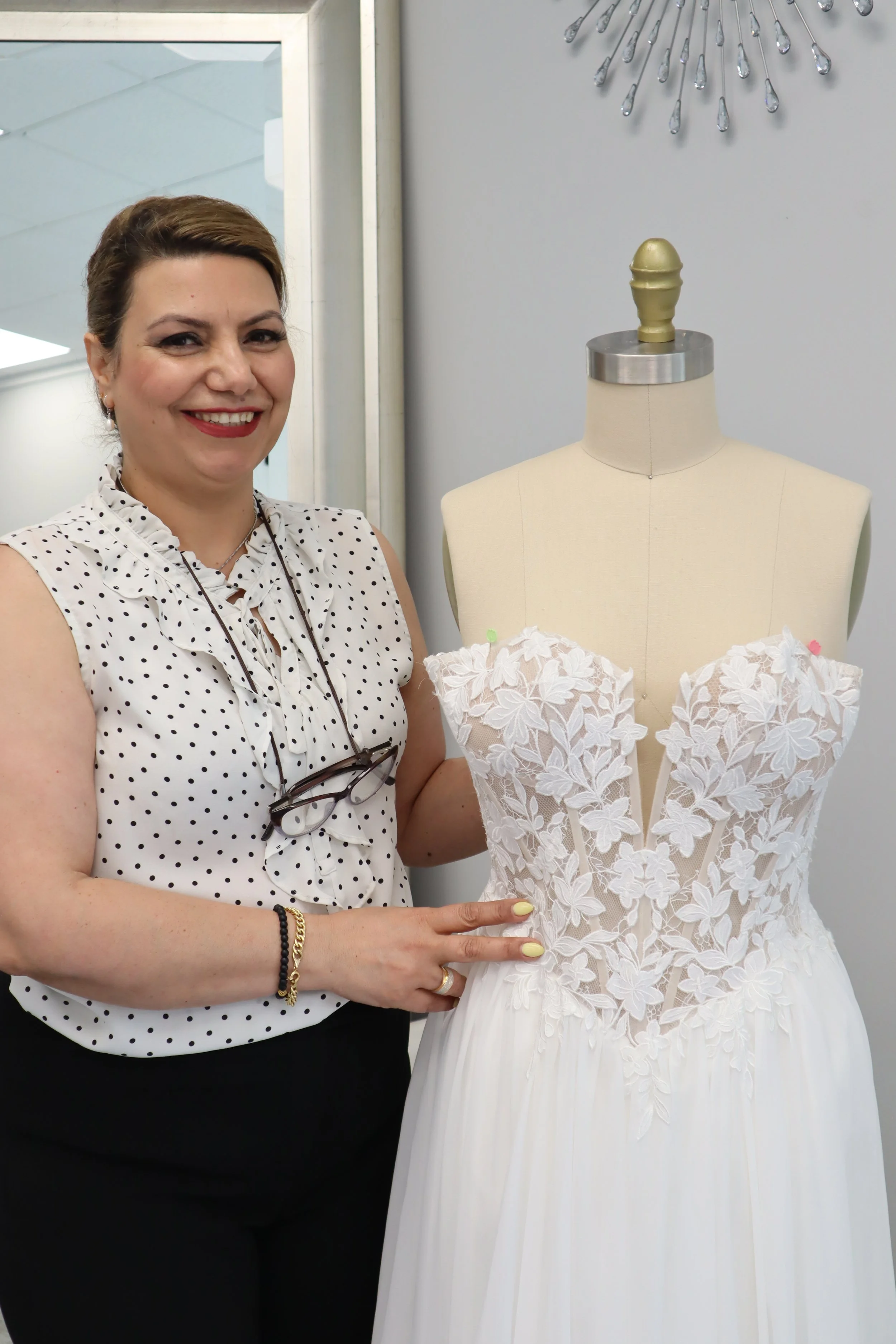 A woman smiling and standing next to a mannequin dressed in a white wedding gown with floral appliqué details, indoors with modern decor.