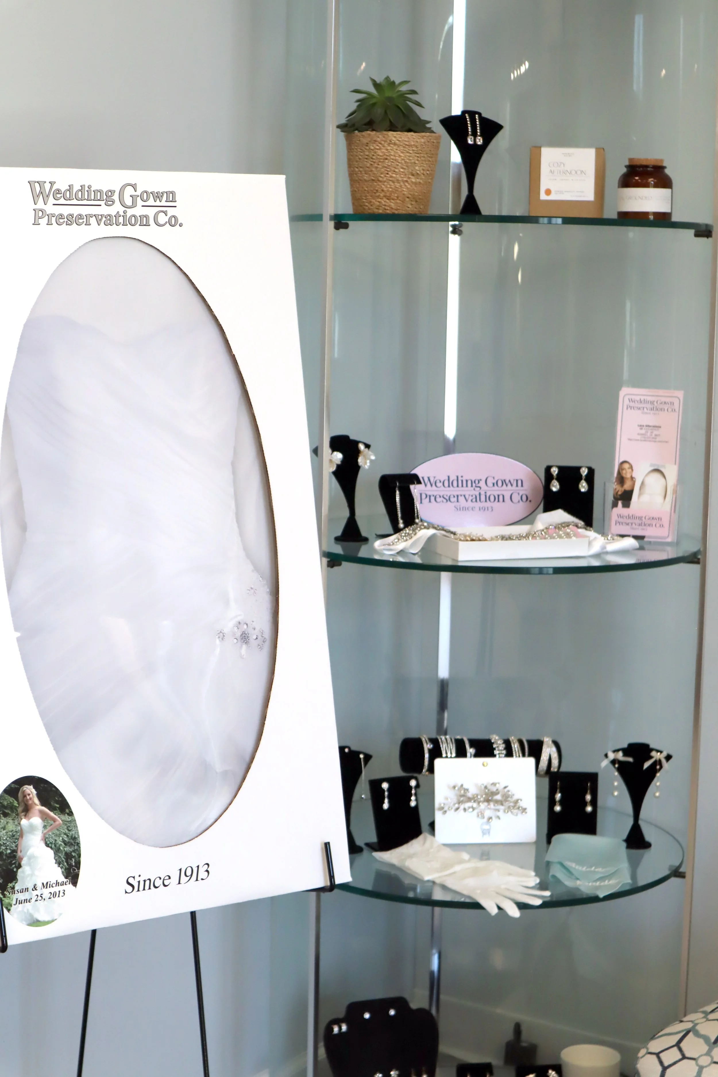 Display of jewelry items, including earrings, bracelets, and necklaces, on black stands and a white surface. A sign indicates it is from Wedding Gown Preservation Co., established in 1913, with a wedding dress poster and an indoor plant on a glass shelf in the background.