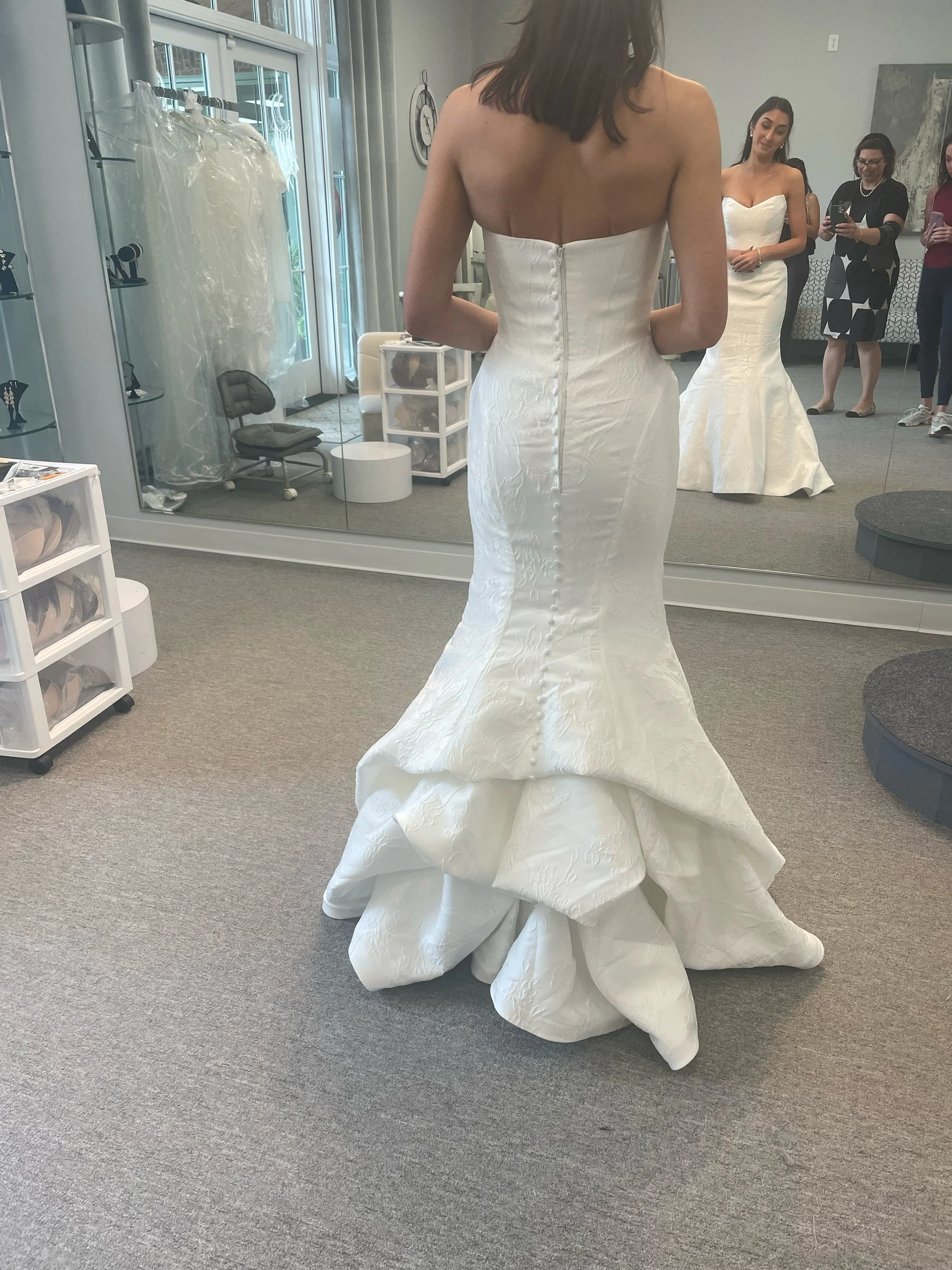 A woman trying on a wedding dress in front of a large mirror, with several people watching, some taking photos, in a bridal boutique