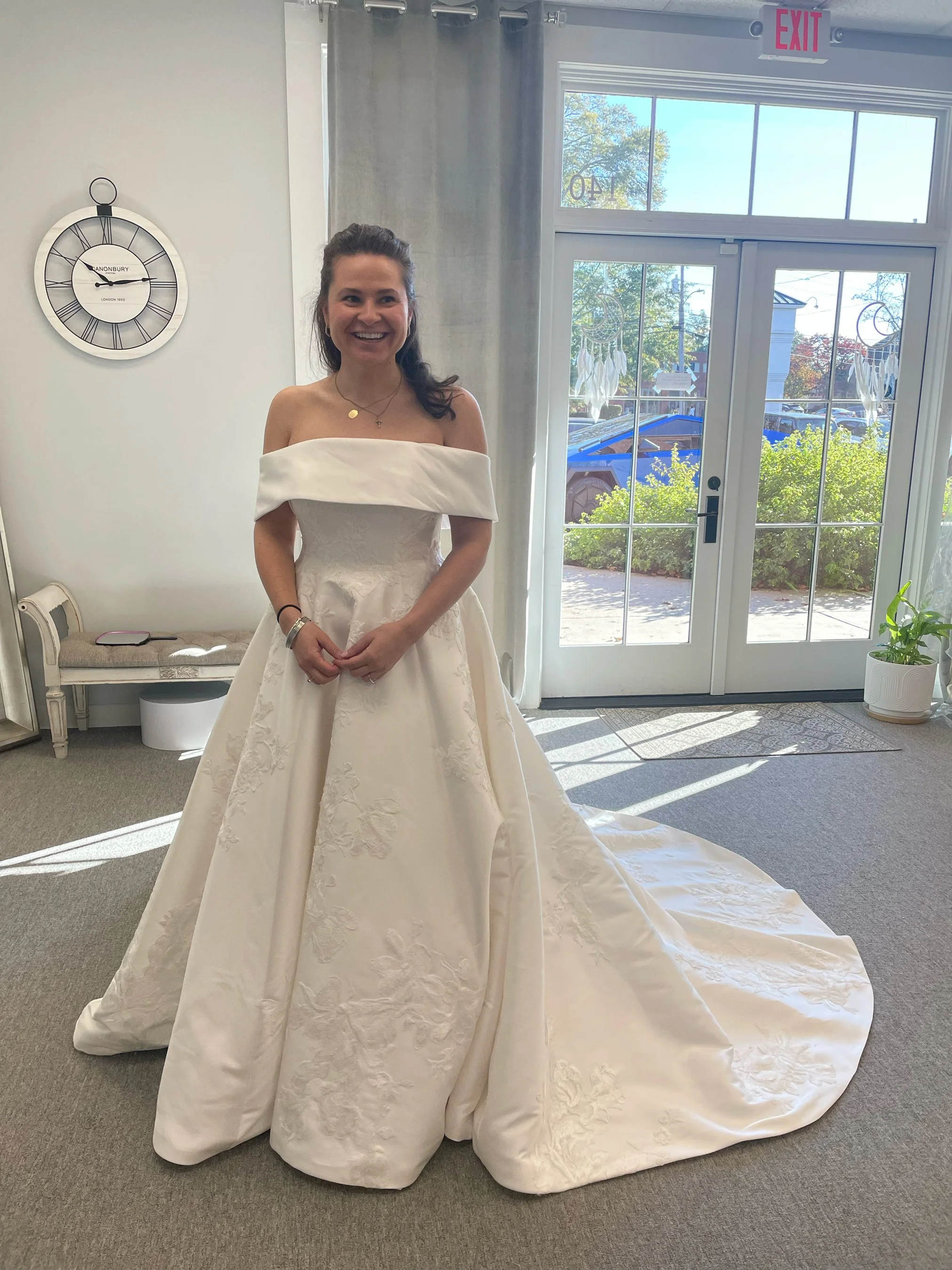 A woman in a white off-shoulder wedding dress standing inside a room with large glass doors, a wall clock, and a bench.
