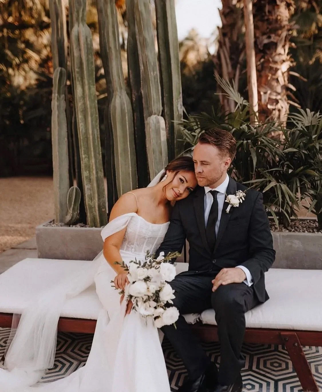 A bride and groom sitting close together on a white cushioned bench outdoors, with the bride resting her head on the groom's shoulder, holding a bouquet of white flowers. They are surrounded by desert plants, including tall cacti and leafy shrubs, in a natural setting during daylight.