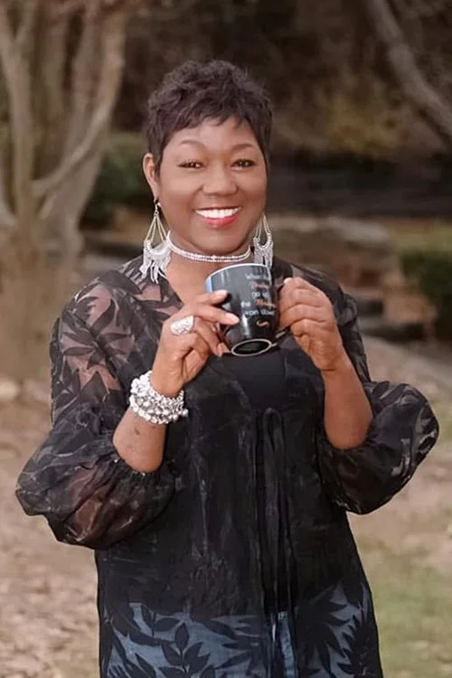 A woman smiling outdoors, holding a black mug, wearing jewelry and a dark patterned jacket, with trees and branches in the background.