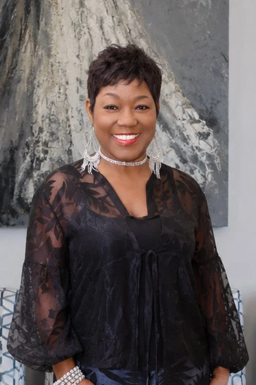 A smiling woman with short dark hair, wearing large earrings, a choker necklace, and a sheer black top with a floral pattern, standing in front of an abstract painting.