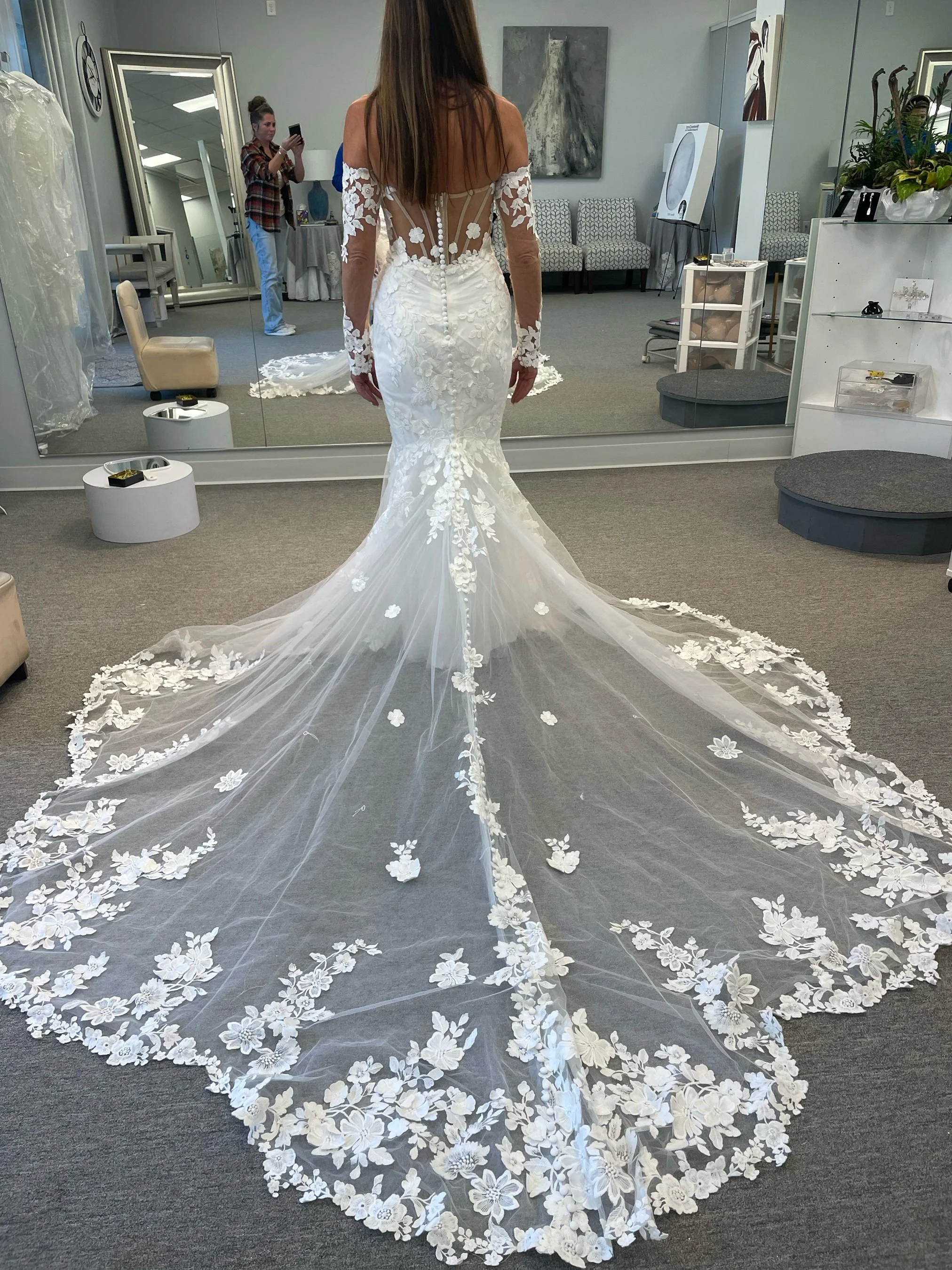 A woman in a wedding dress trying it on in a bridal boutique fitting room, with a large floral train extending behind her.