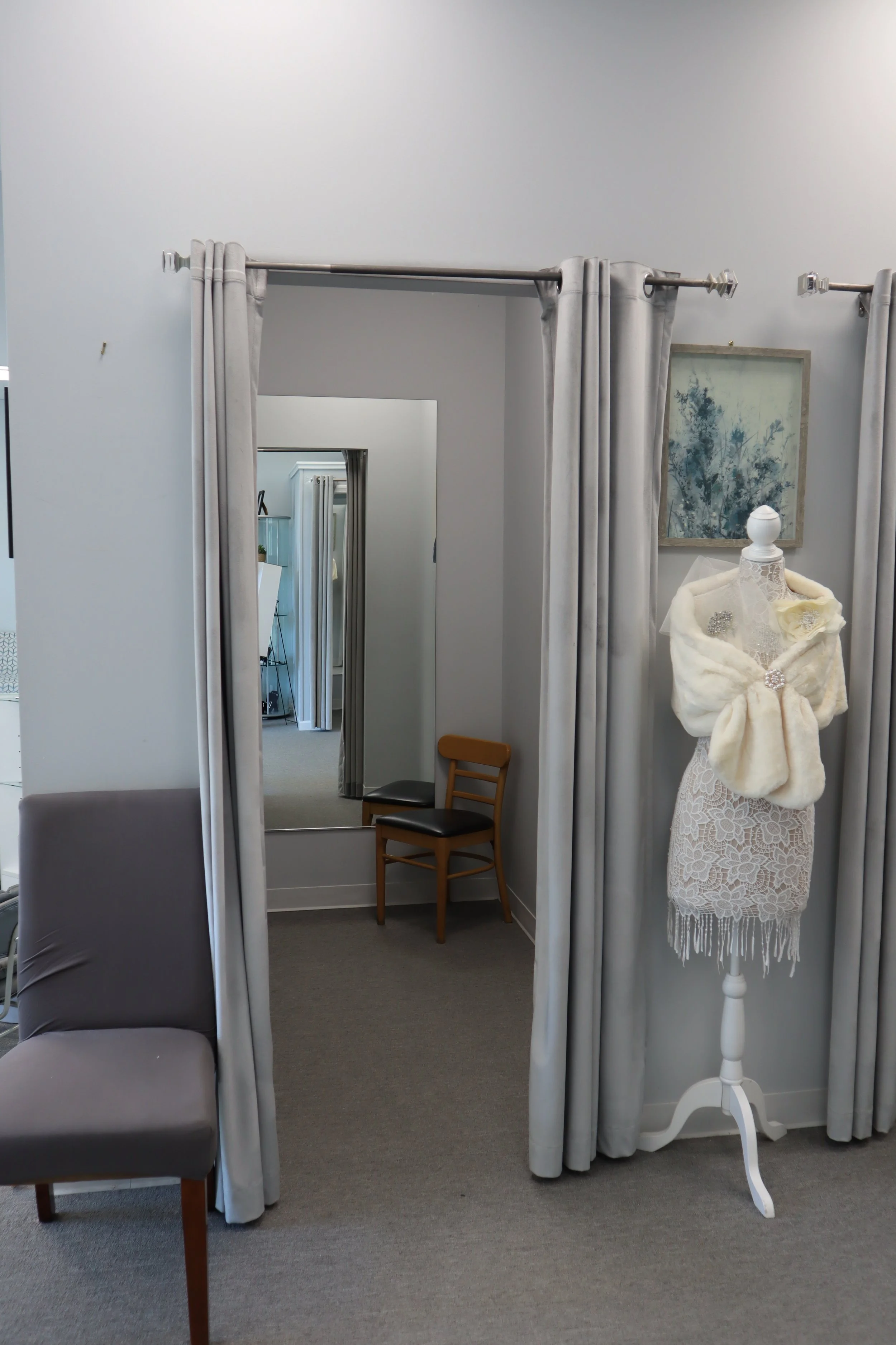 A fitting room with a mirror, a chair, and a dress form with a lace dress and jewelry, framed by curtains.