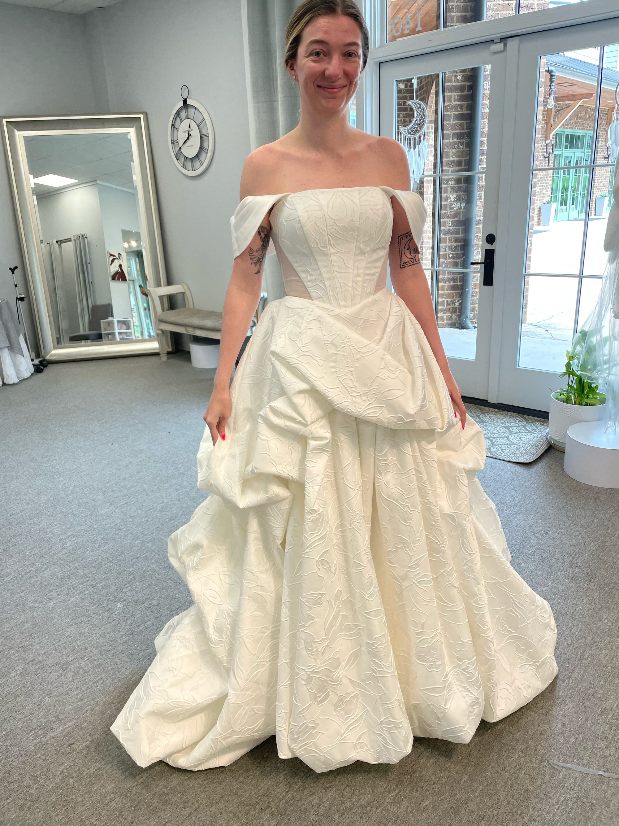 A woman trying on an off-the-shoulder wedding dress in a fitting room, smiling, with tattoos on her arms and a serene indoor setting with a large mirror, wall clock, and glass doors in the background.