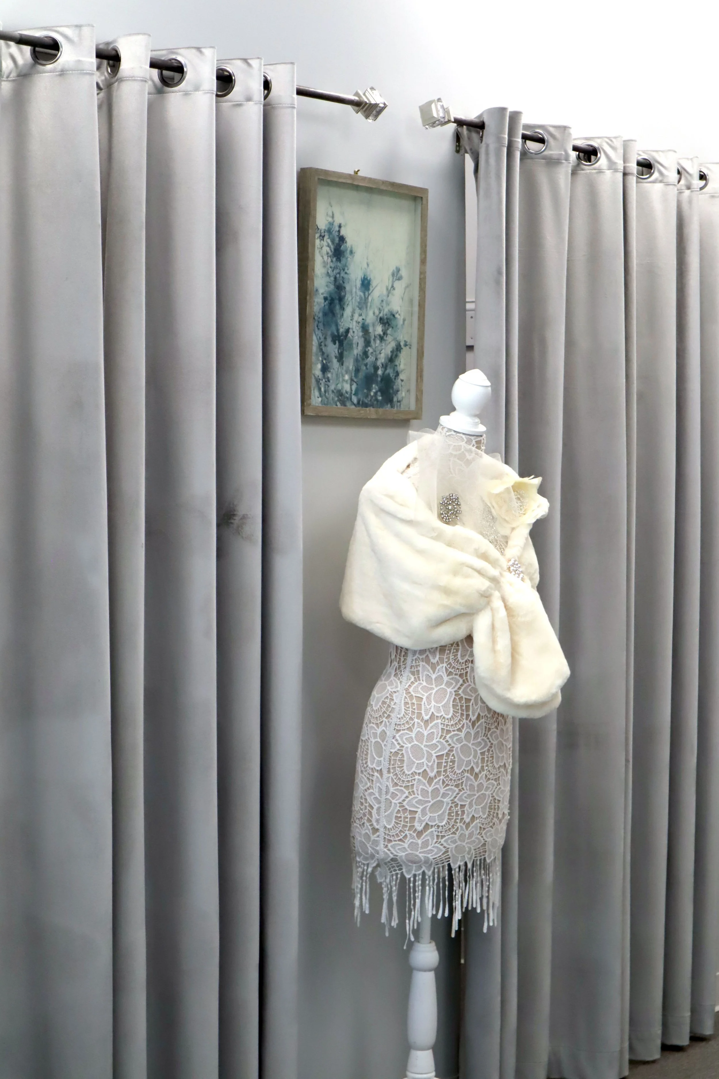 A dress mannequin displays a white lace dress with intricate floral patterns and fringe at the hem. It is accessorized with a cream-colored faux fur stole and brooches. The mannequin is positioned in front of a wall with gray curtains, a framed abstract artwork, and a portion of a white lamp visible.