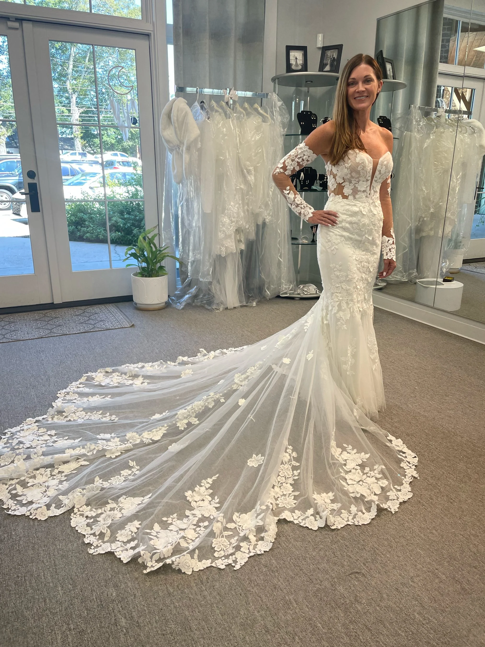 A woman wearing a white wedding dress with a long train and lace details, standing indoors with wedding dresses on racks and a plant in the background.