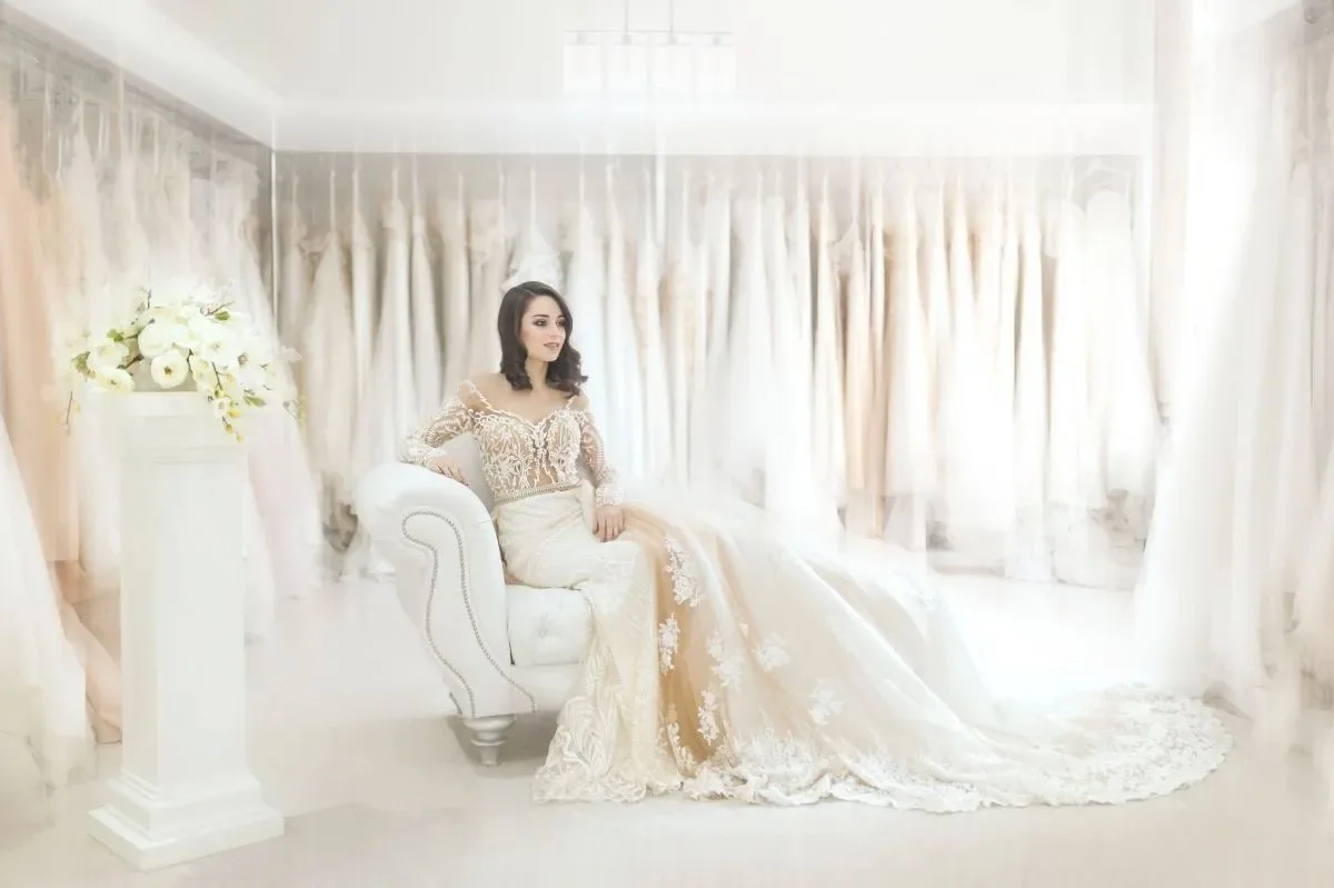 A woman in a wedding dress sitting on a white sofa in a bright room with floral curtains.