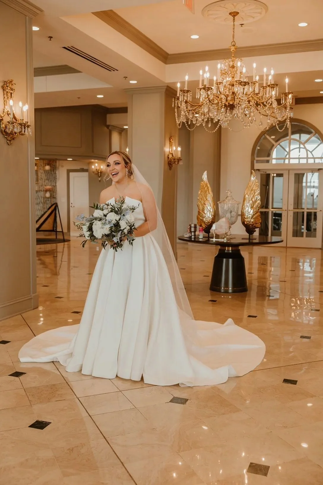 A bride in a white wedding gown holding a bouquet, standing in an elegant, chandelier-lit hotel lobby or reception hall.