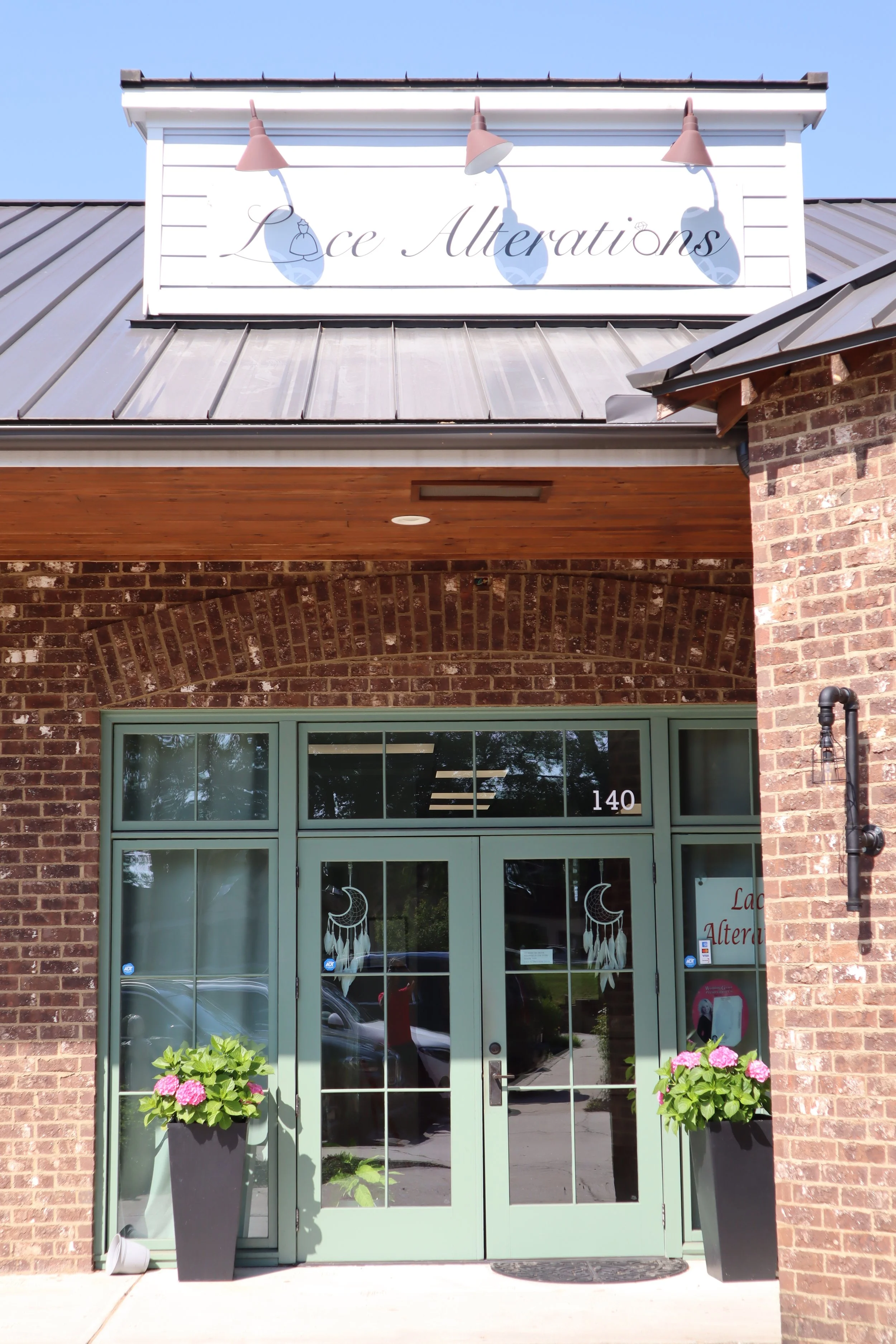 Storefront with a sign reading "Lace Alterations" above the door. The entrance has double glass doors with a green frame, two tall black planters with pink and green flowering plants on either side, and a brick facade. The store number 140 is visible on the door. There are two large windows with lace pattern decorations.