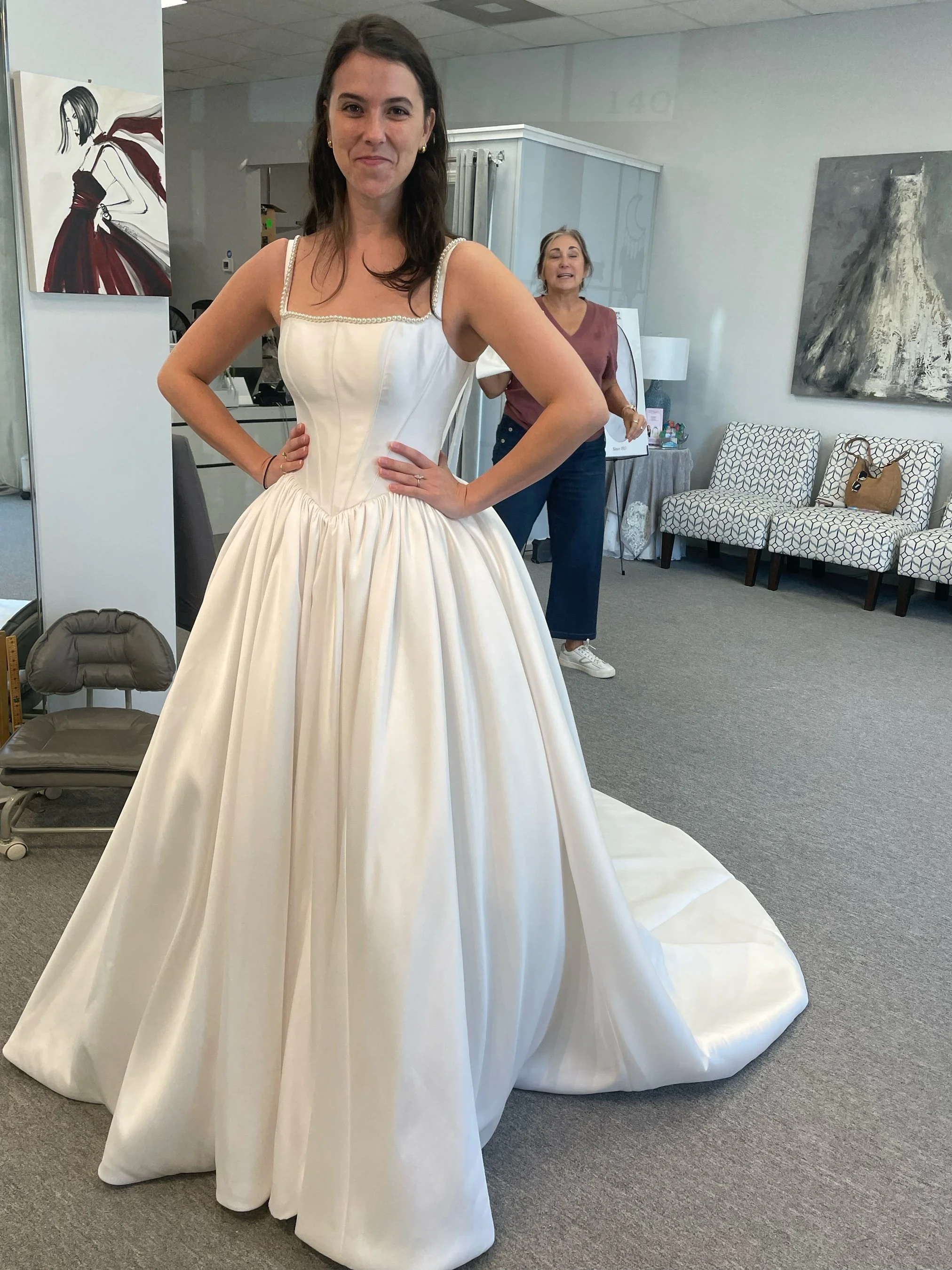 A woman trying on a white wedding gown with a fitted bodice and full skirt, standing in a room with artwork and chairs in the background.