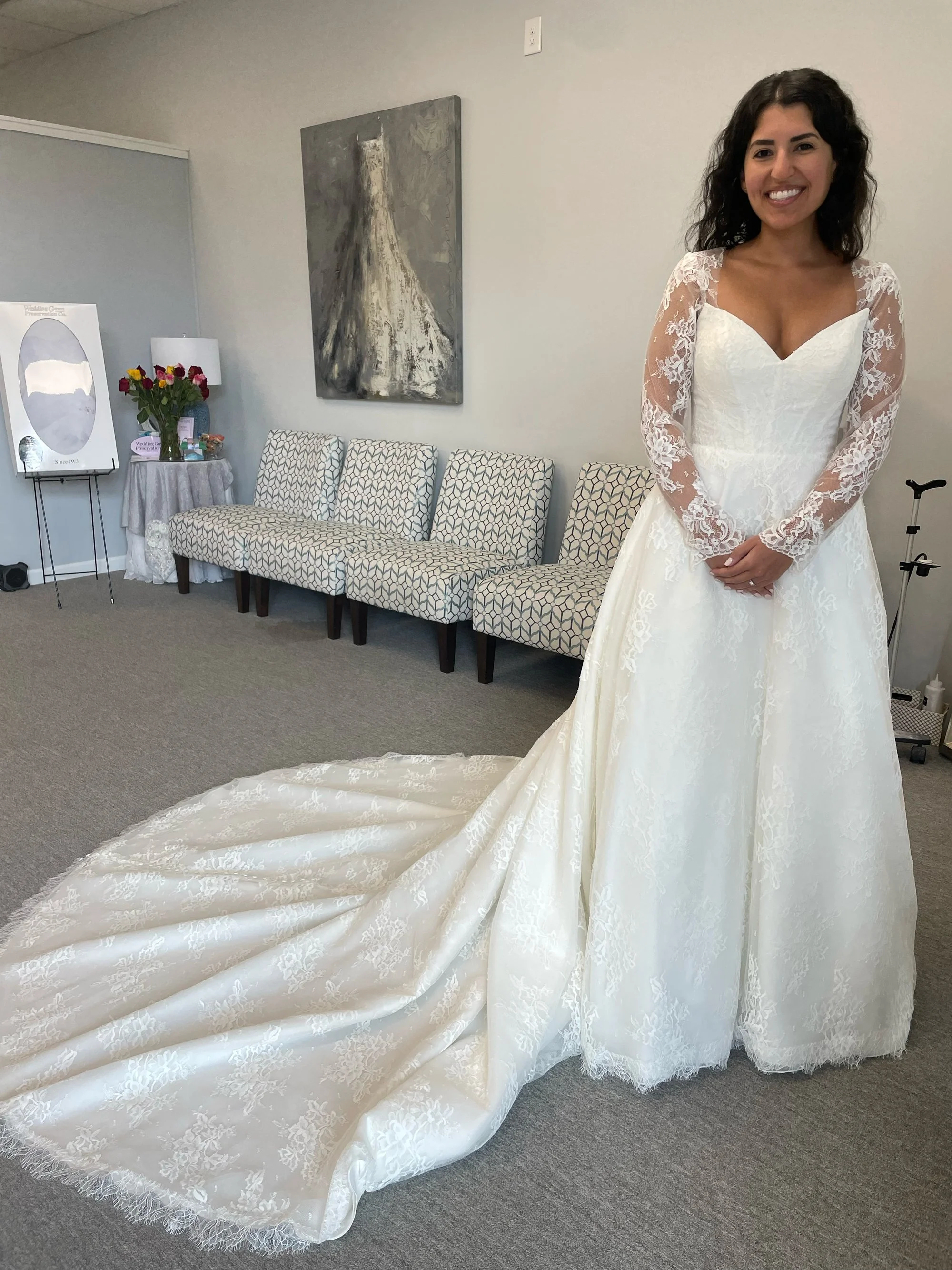 A woman in a wedding gown standing in a room with a floral patterned couch, a table with a vase of roses, and an abstract painting on the wall.