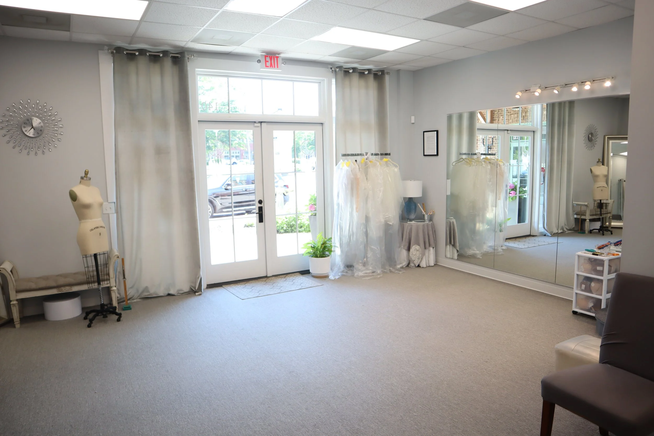 The interior of a small boutique or showroom with large glass doors at the entrance, a mannequin on the left, clothing racks with plastic covers, a table with a lamp, a large mirror on the right wall, and various decorative items.