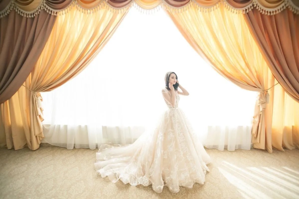 A woman in a white wedding gown standing in front of a large window with golden curtains, backlit by bright light.